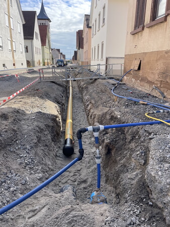 Orangefarbene Gasleitung im Graben und im Vordergrund die blaue provisorische Wasserversorgungsleitung.