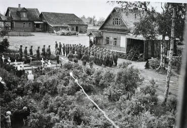 Schwarz-Wei&szlig;foto: Gruppe uniformierter M&auml;nner vor Holzh&auml;usern und einem Friedhof.